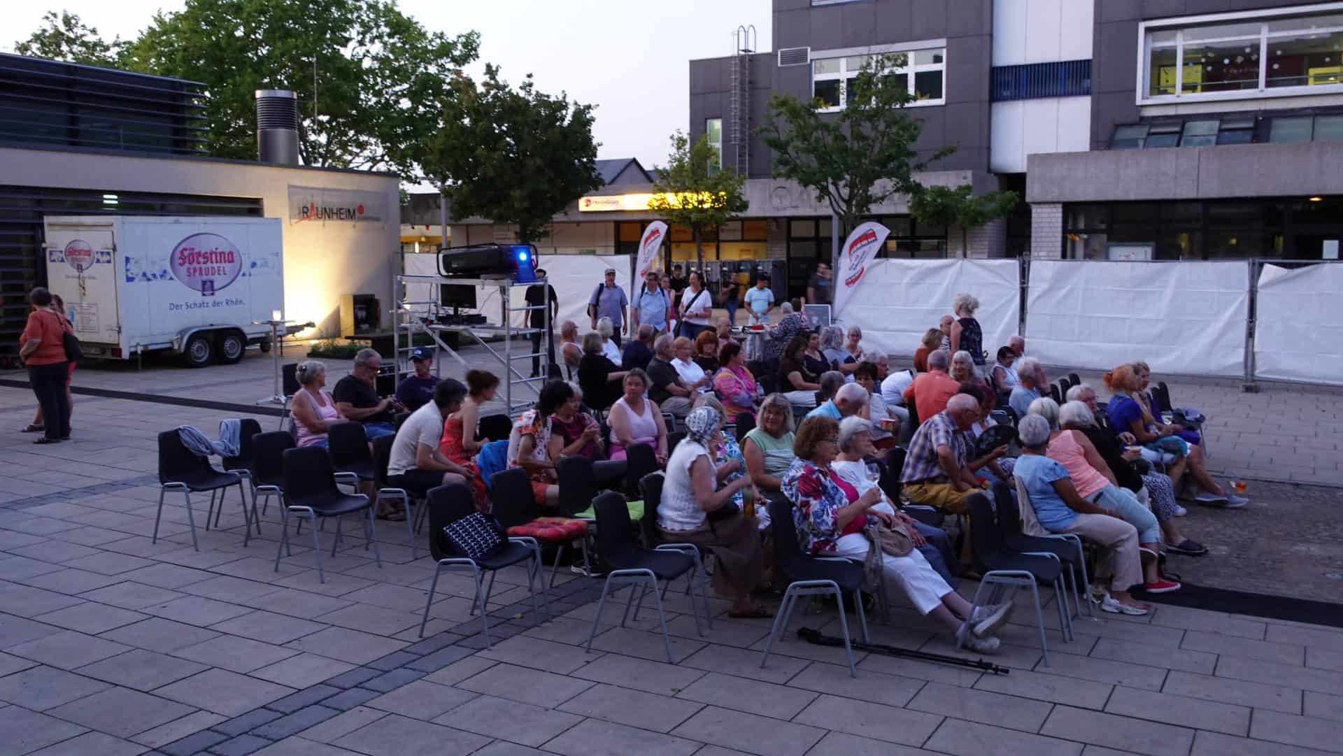 Menschen warten auf Veranstaltung im Freien, Stühle sind aufgestellt.