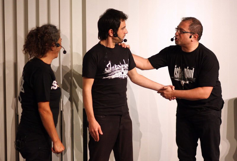 Drei Männer tragen schwarze T-Shirts und sprechen in Mikrofone.