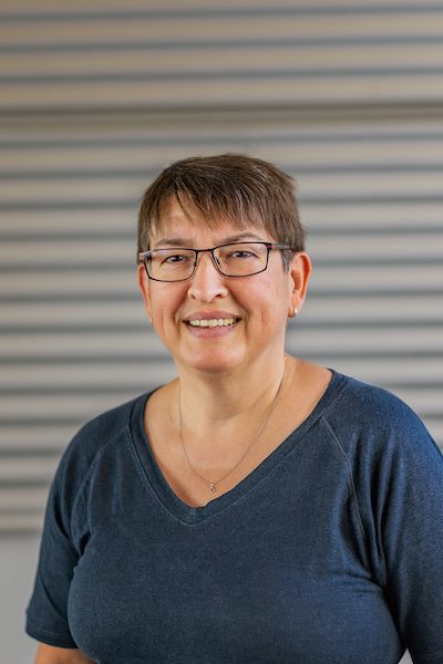 Lächelnde Person mit Brille vor gestreiftem Hintergrund.